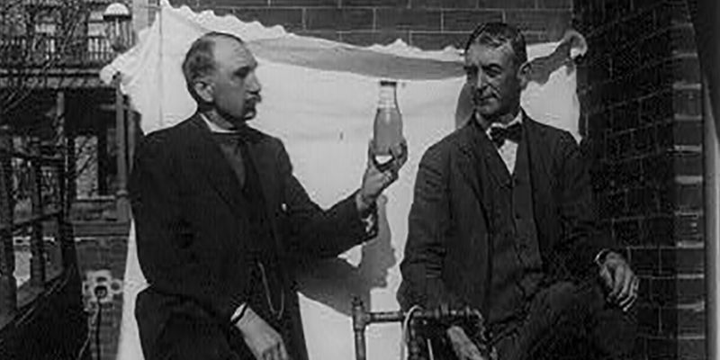 Two men standing outdoors with small still, one of them holding up bottle of liquor